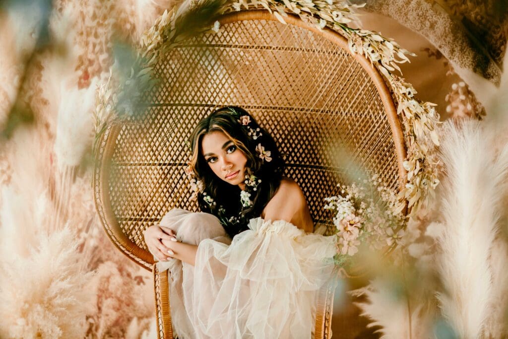 A woman with dark hair has florals added into the hairstyle for decoration as she wears an invory tulle dress and sits curled onto the seat of a wicker peacock chair in front of a boho style photo background with pampas grass in front