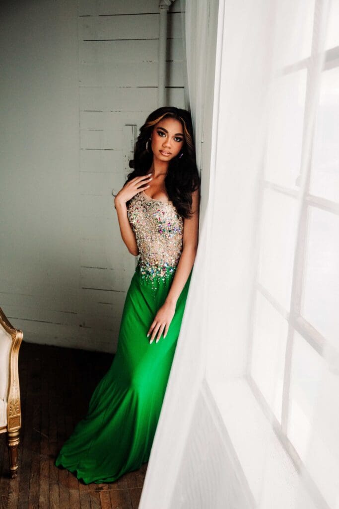 teen girl wears a gown that has silver rhinestones on the top and am emerald green skirt as she leans againt the factory windows and makes eye contact with the camera