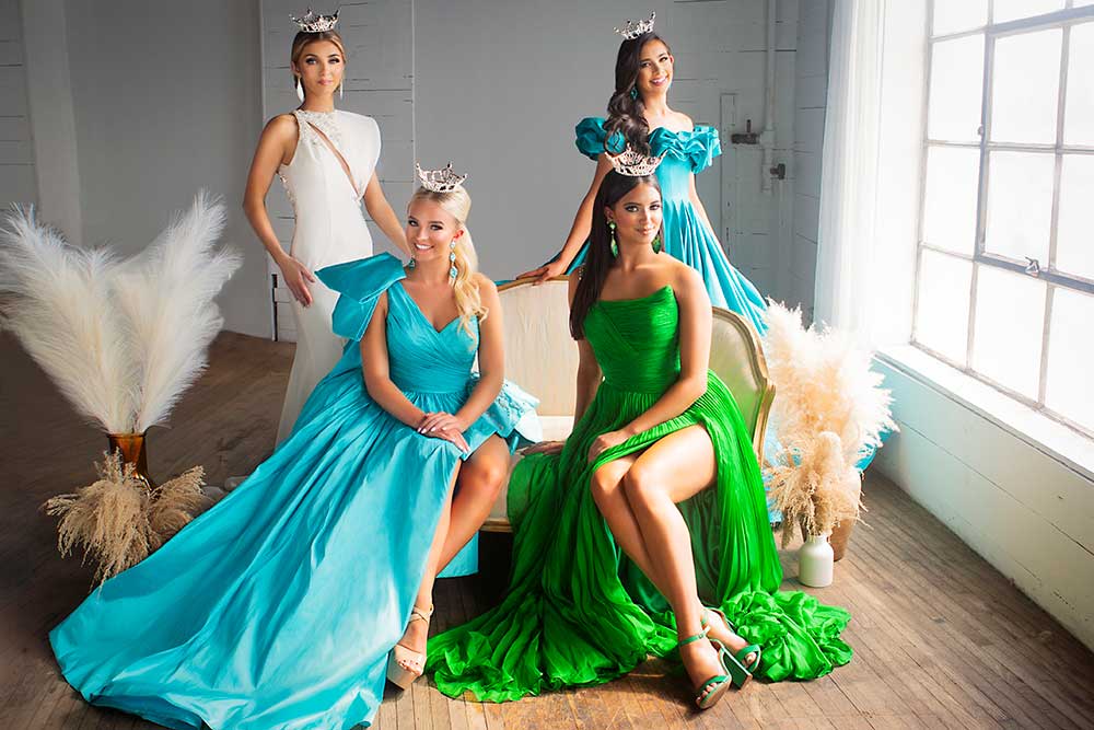 indoor image of 4 women wearing pageant gowns standing side by side (with two sitting on a gold antique sette in the front)
