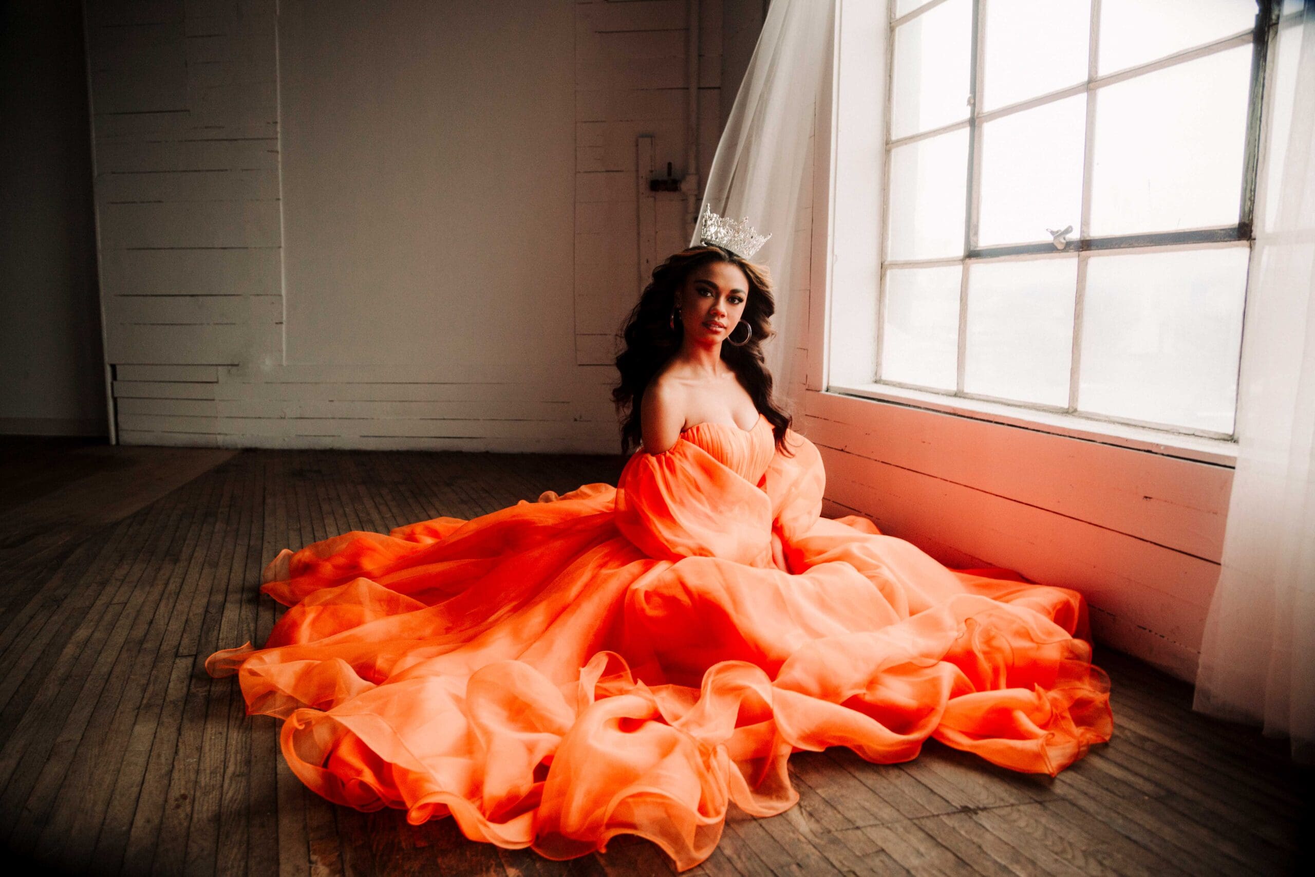 a girl in a big orange gown is sitting on the wooden floor by the factory windows and looking at the viewers with the crown on her head