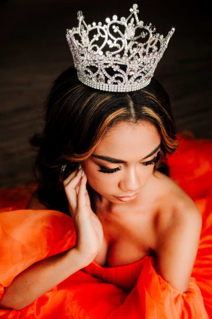 a close up portrait of a girl wearing an orange gown and a large ornate pageant crown. her eyes are looking down
