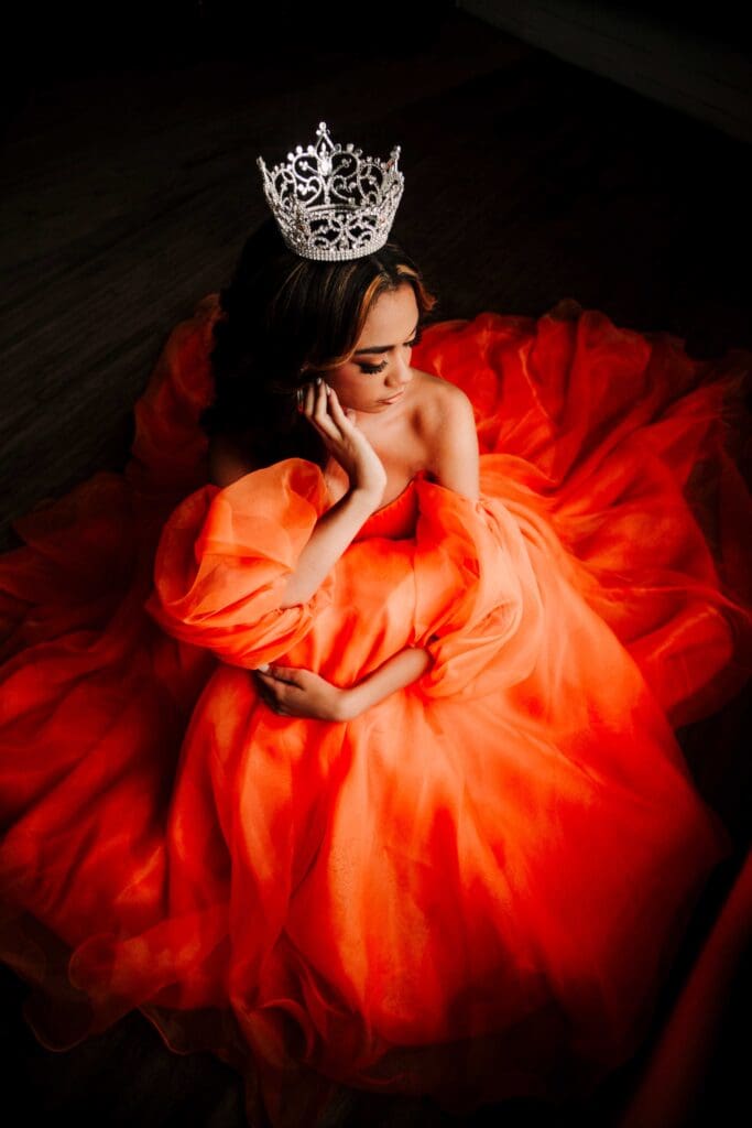 an overhead shot of a teen girl of color sitting on the floor surrounded by her bright orange pageant gown as she hugs her knees and has one hand up to her face, drawing the eye to the silver crown on her head.